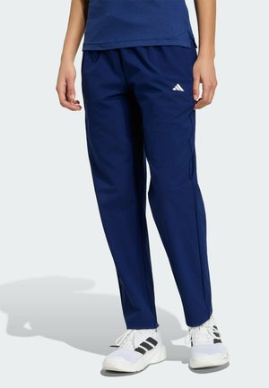 Personne portant un pantalon de survêtement Adidas bleu marine et des baskets blanches Adidas avec des rayures noires, debout sur un fond clair.