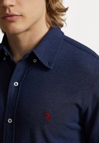 Mörkblå knapp-up skjorta gjord av texturerat tyg med krage, med en liten röd broderad logotyp på bröstet och vita knappar.