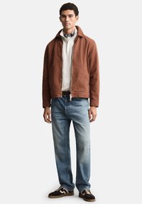 Giacca marrone con zip su un maglione bianco, abbinata a jeans azzurri e sneakers marroni con dettagli bianchi. Design semplice e casual.