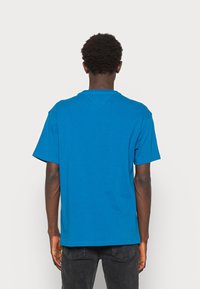 Osoba z krótkimi kręconymi włosami, ubrana w gładką, jasnoniebieską koszulkę i ciemne dżinsy, stojąca tyłem do widza na białym tle.