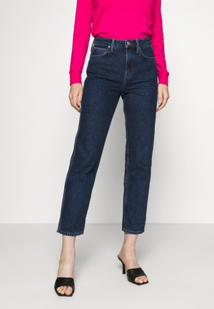 Vrouw draagt donkerblauwe high waist jeans, een felroze top met lange mouwen en zwarte open-teen sandalen met hakken tegen een effen achtergrond.