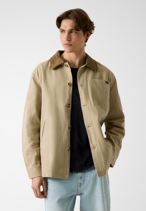 Jeune homme portant une veste beige boutonnée avec un col marron, une chemise noire et un jean bleu clair, debout devant un fond blanc.