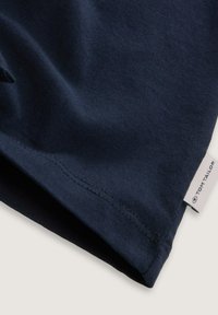 Camiseta de algodón azul marino, textura suave, cuello redondo, mangas cortas, presenta una pequeña etiqueta blanca con "TOM TAILOR" en el dobladillo.