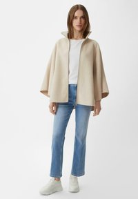 Beżowy płaszcz z wełny z wysokim kołnierzem i zamkiem błyskawicznym z przodu. Połączony z białą koszulką, jasnoniebieskimi dżinsami i białymi sneakerami.