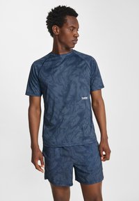 Man i marinblå sportkläder med diskret randmönster, som tittar åt sidan, stående mot en enfärgad vit bakgrund.