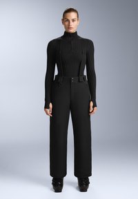 Černý rolák s ribovým texturem a dlouhými rukávy, doplněný černými širokými kalhotami s nastavitelnými popruhy a knoflíkovými detaily.