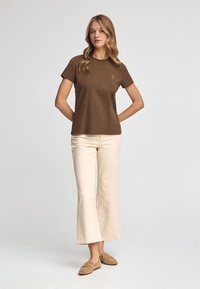 Camiseta de algodón marrón de manga corta con cuello redondo, combinada con pantalones de pierna ancha crema y zapatos de un tono marrón claro sin cordones.