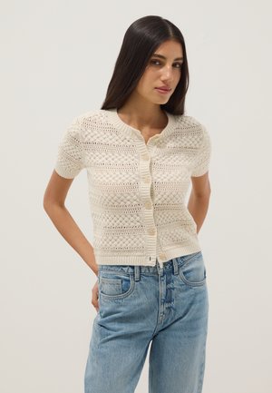 Frau in einem cremefarbenen, kurzärmeligen, gestrickten Cardigan mit Knöpfen, kombiniert mit hellblauen hoch taillierten Jeans, stehend vor einem schlichten Hintergrund.