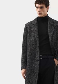 Tmavý plášť s herringbone vzorem, směs vlny, zapínání na jeden knoflík, široké klopy, v kombinaci s černým rolákem a úzkými černými kalhotami.