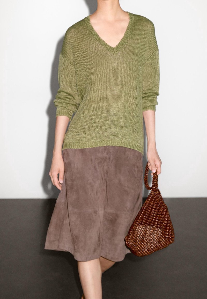 Olijfgroen gebreid sweater met een V-hals, gecombineerd met een bruine suède midi-rok en een gestructureerde bruine kralenhandtas.