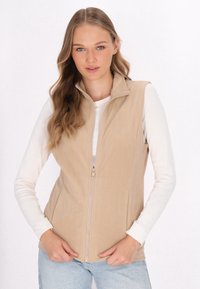 Beige Fleece-Weste mit Stehkragen, vorderem Reißverschluss und seitlichen Taschen. Getragen über einem weißen Langarmshirt, mit glatter Textur.