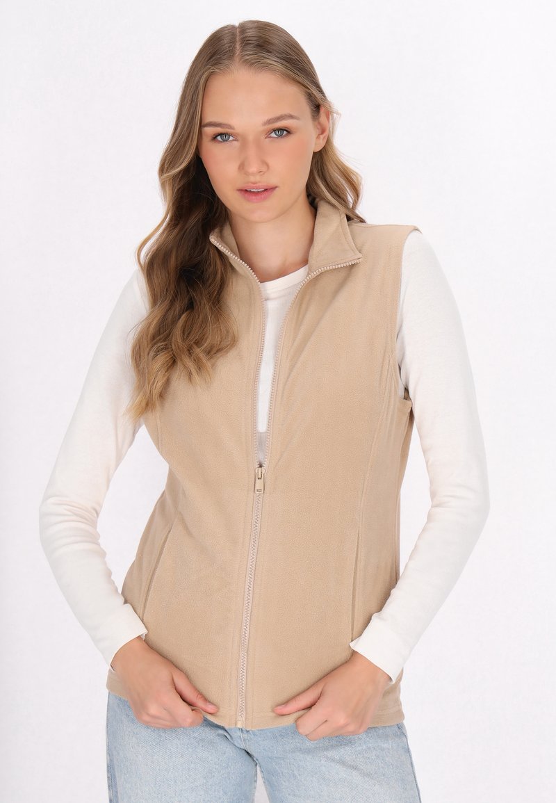 Beige Fleece-Weste mit Stehkragen, vorderem Reißverschluss und seitlichen Taschen. Getragen über einem weißen Langarmshirt, mit glatter Textur.