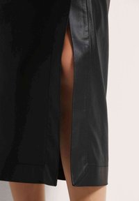 Schwarzer Rock mit hohem Schlitz an der Seite, aus glattem Material mit glänzender Oberfläche und sichtbaren Nähten entlang des Saums.