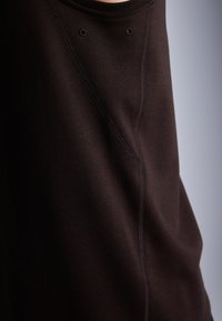 Top de tecido castanho com uma textura suave, decote arredondado e costura visível. Dois pequenos olhos estão posicionados perto do decote.