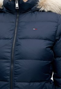 Doudoune bleu marine avec fermeture éclair à l'avant, capuche bordée de fourrure, et petit logo brodé rouge, blanc et bleu sur la poitrine.