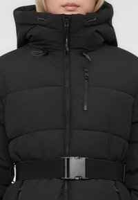 Άτομο που φοράει ένα μαύρο επενδυμένο μπουφάν με ψηλό γιακά, κουκούλα, τσέπη με φερμουάρ στο στήθος και ρυθμιζόμενη μαύρη ζώνη στη μέση με πλαστικό κούμπωμα.