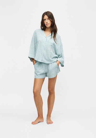 Femme aux longs cheveux portant une chemise boutonnée ample rayée bleu clair et blanc et un short assorti, debout pieds nus les mains dans les poches.