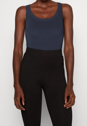 Femme portant un haut bleu marine sans manches et un pantalon noir taille haute, debout devant un fond clair uni.