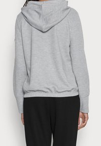 Person in einem hellgrauen Kapuzenpullover mit elastischem Bund und schwarzen Hosen, von hinten vor einem einfarbigen Hintergrund gezeigt.