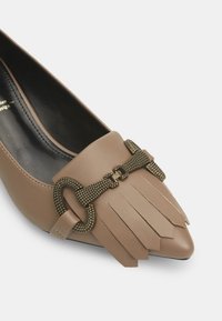 Scarpa in pelle beige con punta affusolata, dettagli in frange e una fibbia decorativa in bronzo sulla tomaia. Texture liscia e tacco basso.