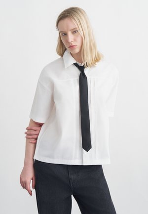 Mladá žena v bielej krátkoročnej košeli s čiernou kravatou a tmavými nohavicami stojaca pred jednoduchým bielym pozadím.