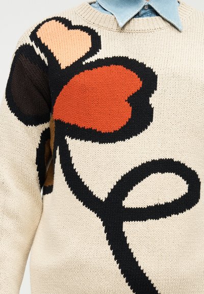 Κρεμ πλεκτό πουλόβερ με μεγάλο σχέδιο λουλουδιού με μαύρο κοτσάνι και κόκκινα, ροδακινί και καφέ πέταλα σε σχήμα καρδιάς, φορεμένο πάνω από μπλε πουκάμισο με γιακά.