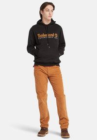 Felpa nera con cappuccio e logo "Timberland" arancione, abbinata a pantaloni in corduroy marroni e stivali marroni con lacci.