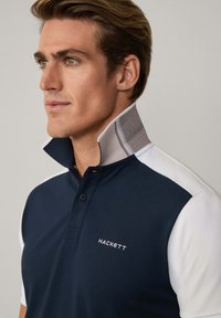 Polo de color azul marino con una sección blanca en el hombro. Presenta un cuello abotonado con un interior texturizado. Logo "HACKETT" bordado en la parte delantera.