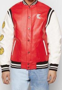 Röd läder-varsityjacka med vita läderärmar som har gula leende ansikten, ribbade muddar, krage och midja med svarta ränder.