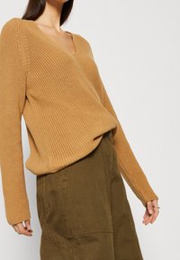 Maglione a V in colore beige con una texture a coste, abbinato a pantaloni larghi verde oliva con tasche e dettagli di cucitura.