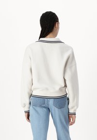 Persoană cu părul împletit, purtând un pulover alb cu margini negre și blugi de culoare albastru deschis, stând cu spatele la un fundal alb.