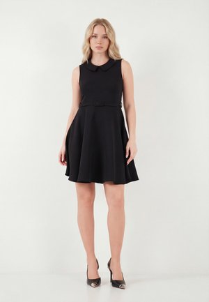 Žena stojaca v čiernych šatách bez rukávov s golierom a opaskom, kombinovaných s čiernymi lodičkami na bielom pozadí.