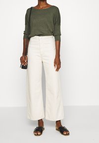 Olivgrön stickad tröja med vida ärmar, kombinerad med krämfärgade vida byxor. Modellen bär svarta sandaler och håller en liten svart axelväska.