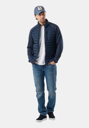 Mand iført marineblå quiltet jakke, hvid t-shirt, blå jeans, sorte sneakers og grå baseballkasket med "Go Coco"-mærke, stående med hænderne i lommerne.