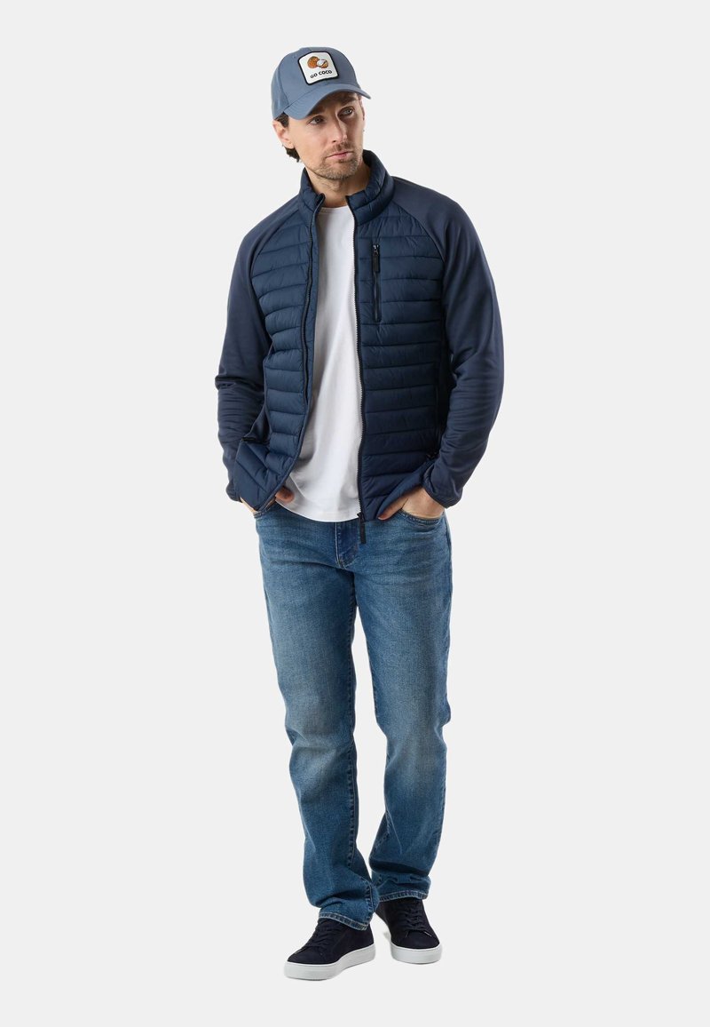 Mężczyzna w granatowej pikowanej kurtce, białej koszulce, niebieskich dżinsach, czarnych trampkach i szarej czapce z daszkiem z naszywką "Go Coco", stojący z rękami w kieszeniach.