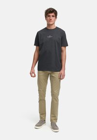 Mørk grå t-skjorte med korte ermer og hvit logo, kombinert med lyse chinos og grå sko, stående på en ensfarget bakgrunn.