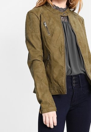 Femme portant une veste zippée vert olive sur un chemisier gris foncé avec un col en dentelle, associée à un pantalon taille haute foncé.