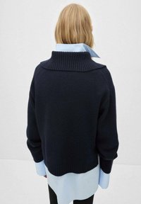 Blonde person facing away wearing a navy blue ribbed sweater layered over a light blue shirt with an upturned collar.