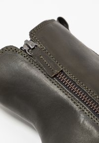 Botte en cuir vert olive avec une fermeture éclair métallique et des surpiqûres contrastées. Le design présente une texture lisse et une forme robuste.