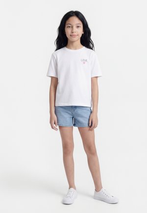 Ung pige med langt mørkt hår iført en hvid "LOVE" T-shirt, denimshorts og hvide sneakers, stående foran en ensfarvet hvid baggrund.