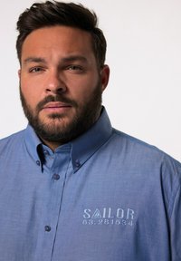 Camicia a maniche lunghe di colore azzurro chiaro con colletto, caratterizzata da ricami con la scritta "SAILOR" e dettagli numerici. Tessuto morbido con una consistenza liscia e bottoni piccoli.
