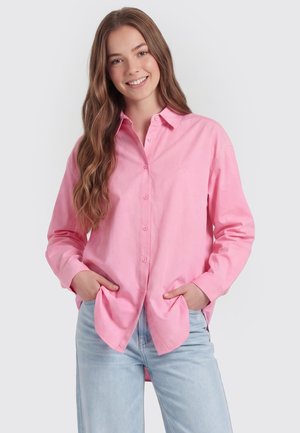 Junge Frau lächelt mit langen, welligen braunen Haaren, trägt ein lockeres rosa Hemd mit Knöpfen und hellblaue Jeans, die Hände in den Taschen.