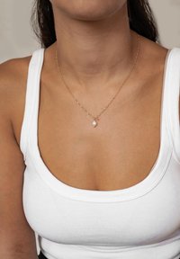 Złoty naszyjnik z zawieszką w kształcie łzy, noszony na prążkowanym białym topie na ramiączkach. Gładka faktura z delikatnymi detalami łańcuszka.