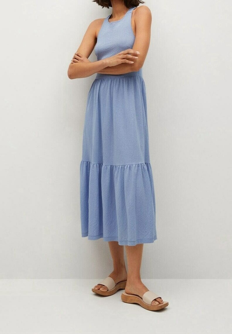 Femme portant une robe midi bleu clair sans manches avec un volant froncé et des sandales beige à enfiler, bras croisés.