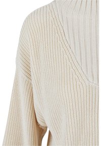 Krémově barevný pletený svetr s žebrovanou strukturou, hlubokým výstřihem do V a širokými rukávy. Celý svetr zdobí jemný vertikální vzor.