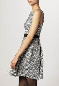 Abito a fantasia bianco e nero con corpino aderente, gonna svasata e cintura nera, dotato di spalline sottili e una consistenza generale liscia.