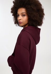 Felpa bordeaux in tessuto morbido con vestibilità rilassata. Presenta un cappuccio e una tasca a marsupio, evidenziando le cuciture a vista e una texture liscia.