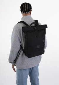 Svart rulltoppsryggsäck med vadderade axelremmar, utrustad med en framficka med dragkedja och en stilren design. Materialet är slätt och hållbart.