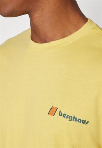 Žluté bavlněné tričko s kulatým výstřihem. Na levé části hrudi je zelené logo "Berghaus" a oranžový akcent. Hladká textura.