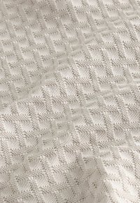 Pletená textilie ve světle béžové barvě s geometrickým vzorem, který obsahuje vystouplé diamantové tvary a má jemný, texturovaný vzhled.